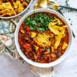 White ceramic bowl filled with beans and chili and cheese on top. Gold and black spoons to the side.