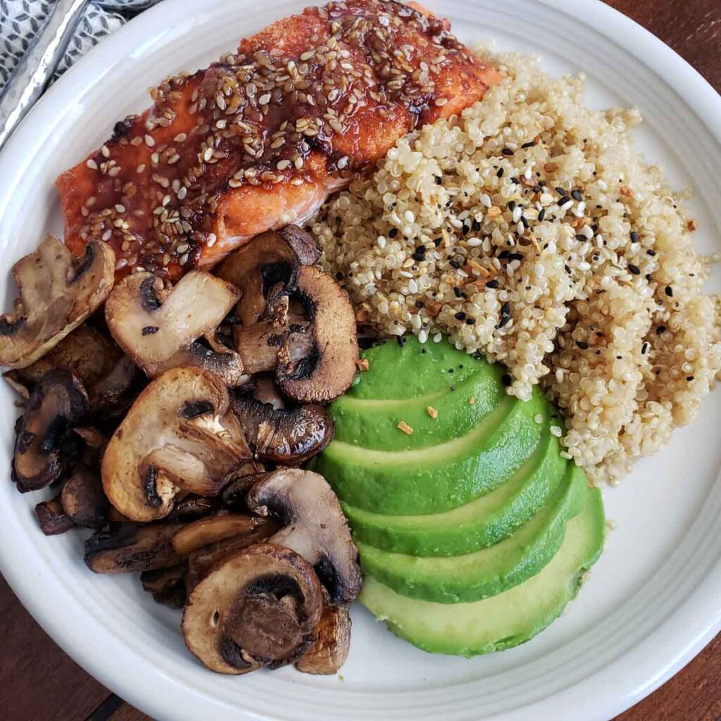 White plate with air fried salmon with teriyaki sauce, quinoa, avocado and mushrooms. 