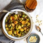 White bowl filled with brussel sprouts and sauce and a silver spoon with sauce. Peanuts on the side.