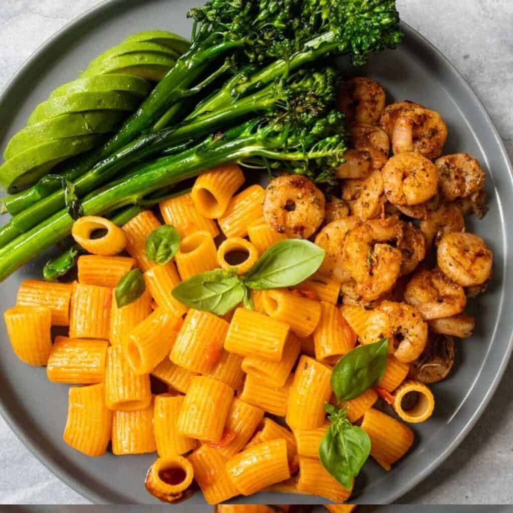 Shrimp and harissa pasta on plate with veggies and avocado. 