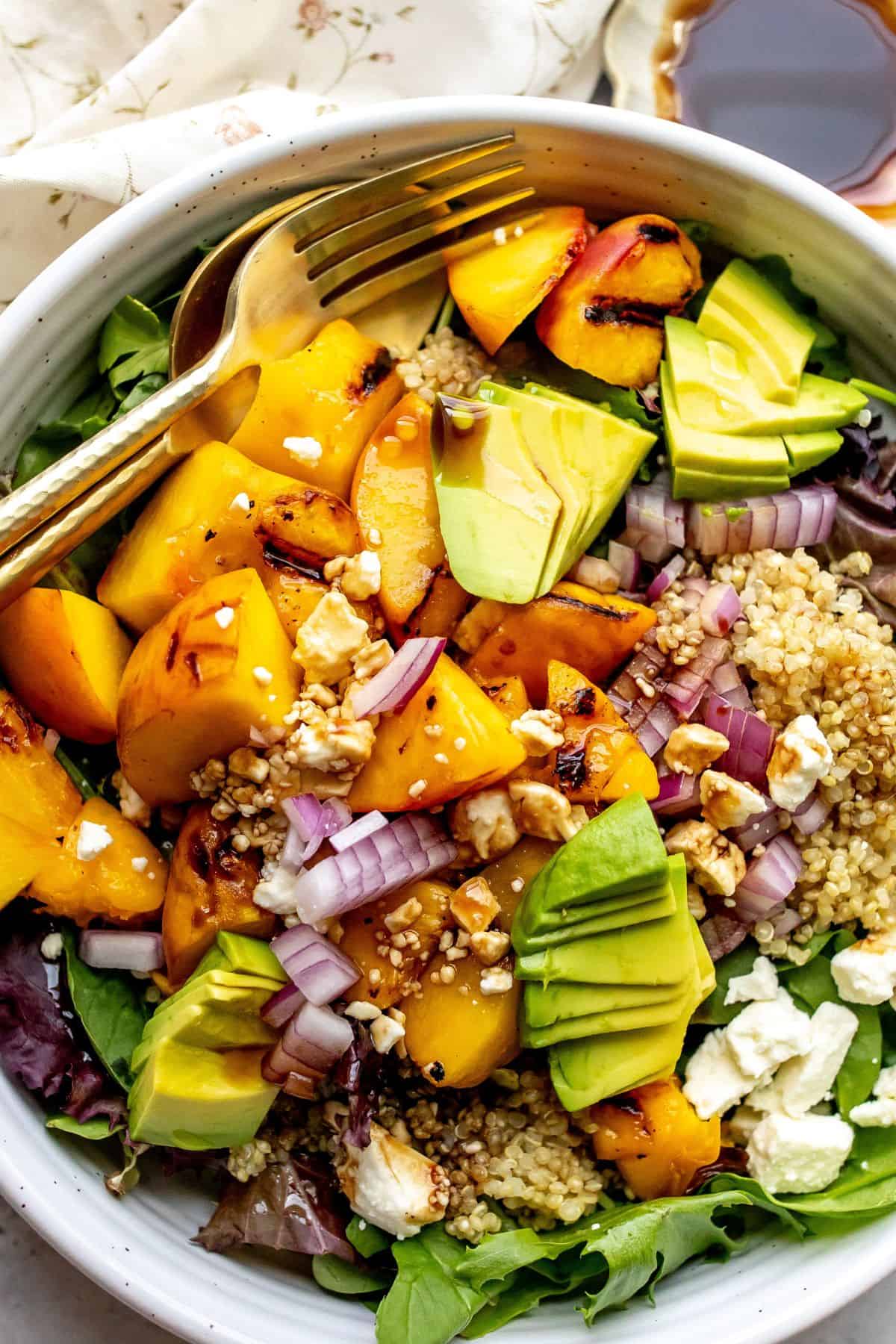 White bowl with peach feta salad and avocado on it, serving utensils to the side.