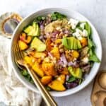 White bowl with peach feta salad and avocado on it, serving utensils to the side.