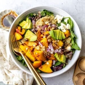 White bowl with peach feta salad and avocado on it, serving utensils to the side.