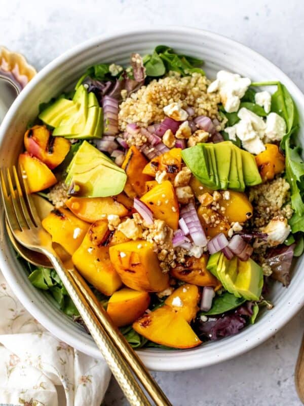 White bowl with peach feta salad and avocado on it, serving utensils to the side.