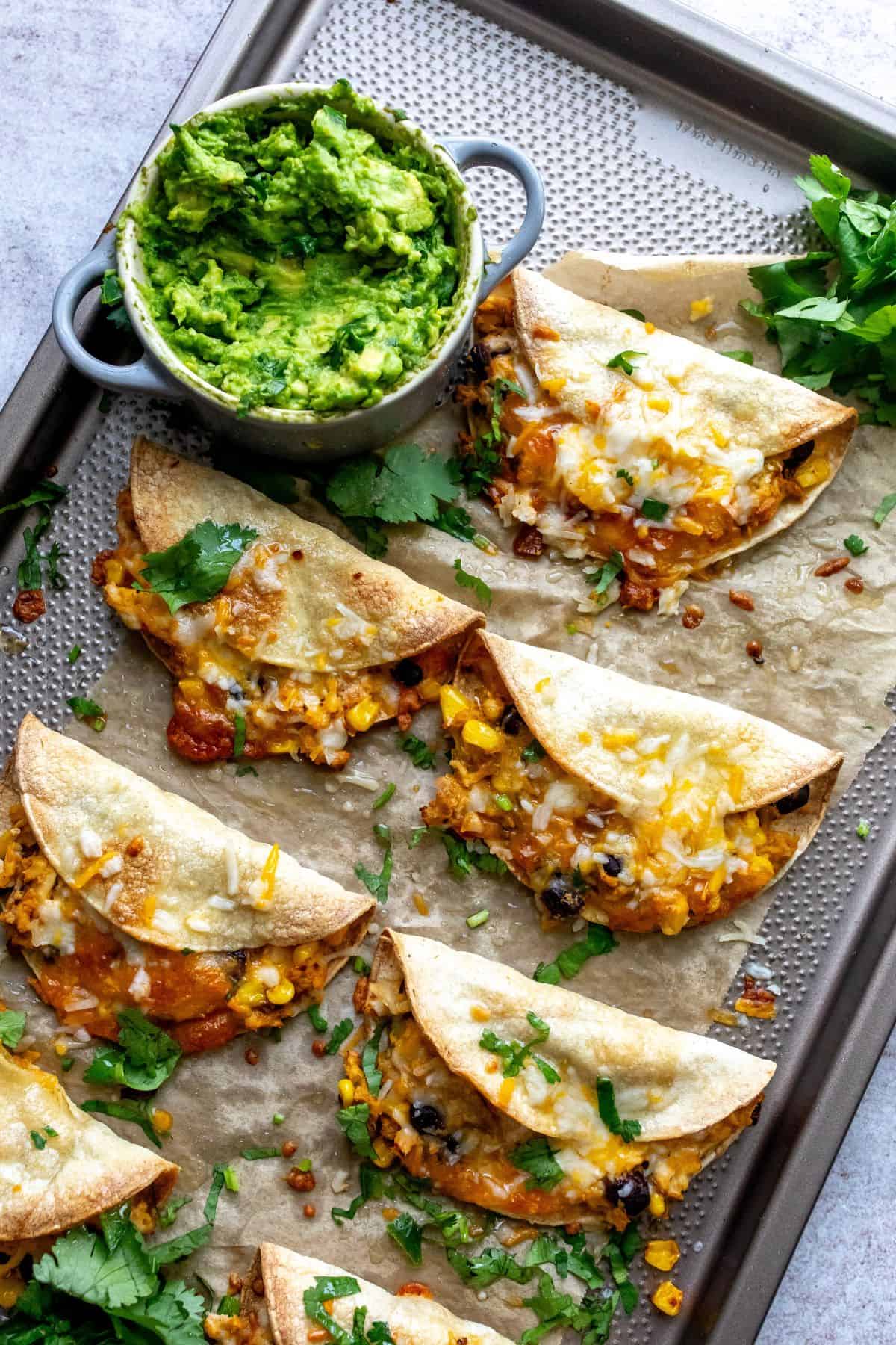 Crispy chicken tacos on a baking sheet with cilantro and guacamole on the side. 