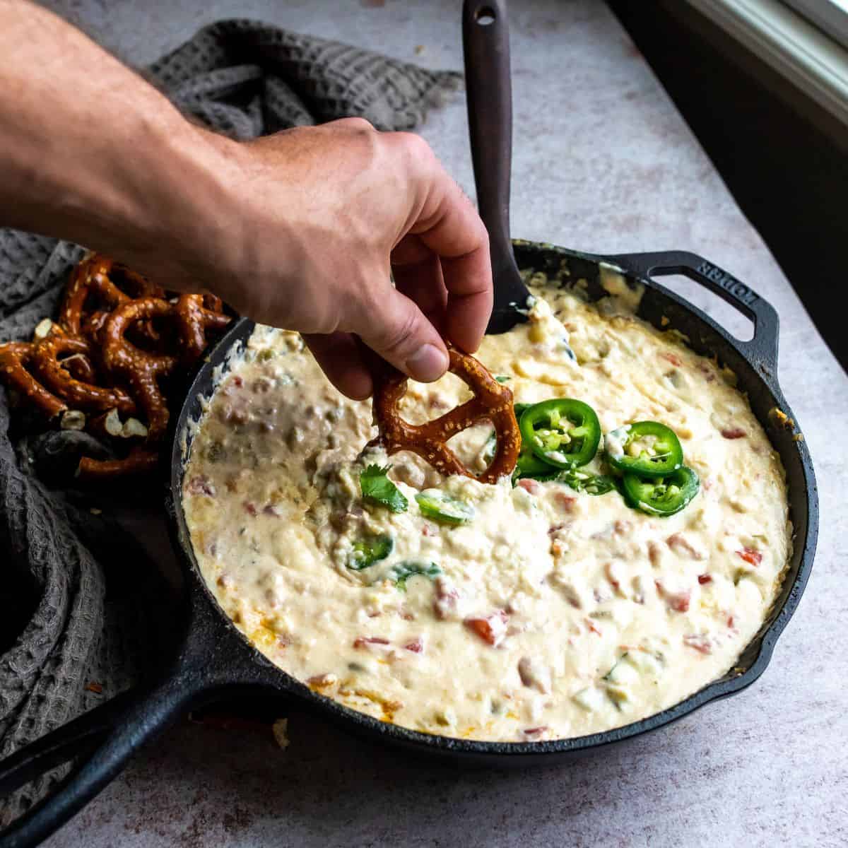 Queso in a cast iron with a hand dipping a pretzel. 