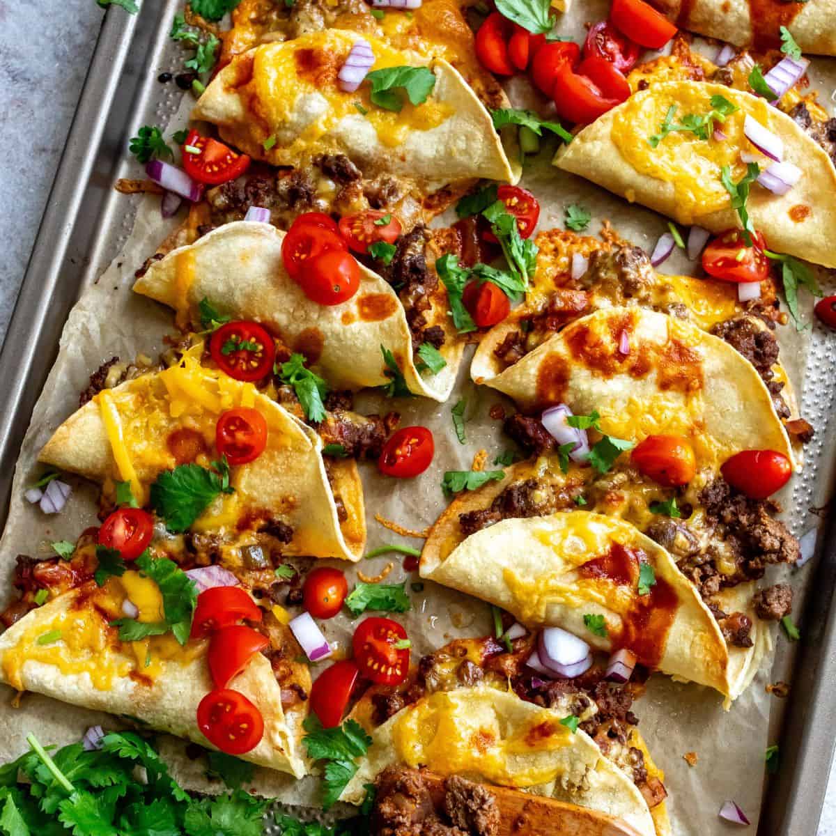 Crispy Beef Tacos on parchment liked baking sheet with hot sauce and tomatoes.