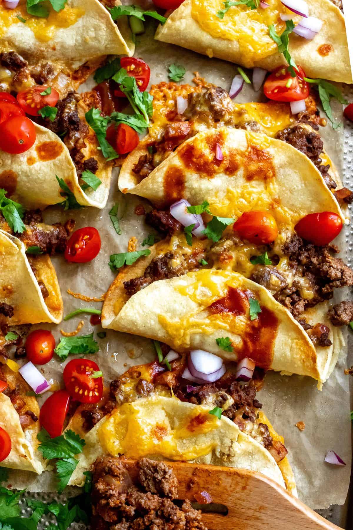 Crispy Beef Tacos on parchment liked baking sheet with hot sauce and tomatoes. 