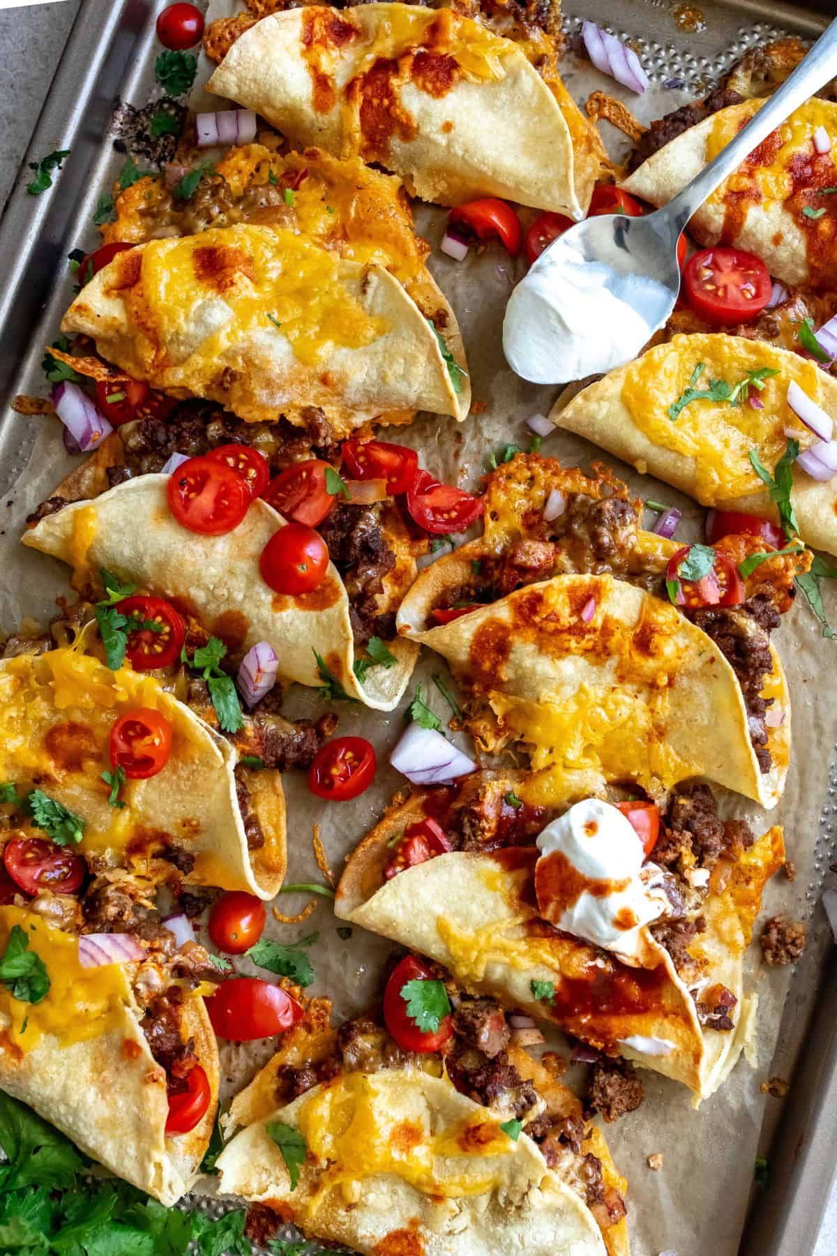 Crispy beef tacos on a sheet pan with sour cream.