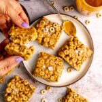 peanut butter and cheerio bars on a white plate with peanut butter on a spoon.