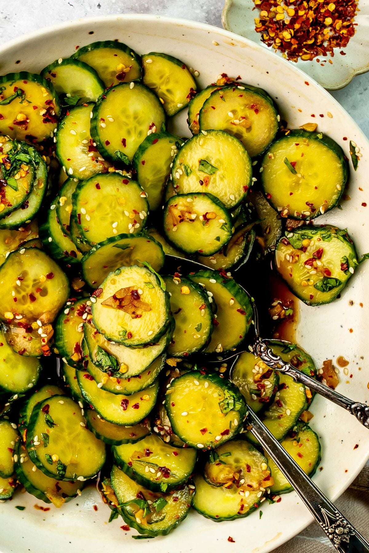 A serving spoon scoops some Asian spicy cucumber salad from a white bowl on a countertop.