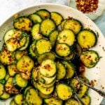 A serving spoon scoops some Asian spicy cucumber salad from a white bowl on a countertop. A small bowl of red pepper flakes sits off to the side.