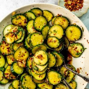 A serving spoon scoops some Asian spicy cucumber salad from a white bowl on a countertop. A small bowl of red pepper flakes sits off to the side.