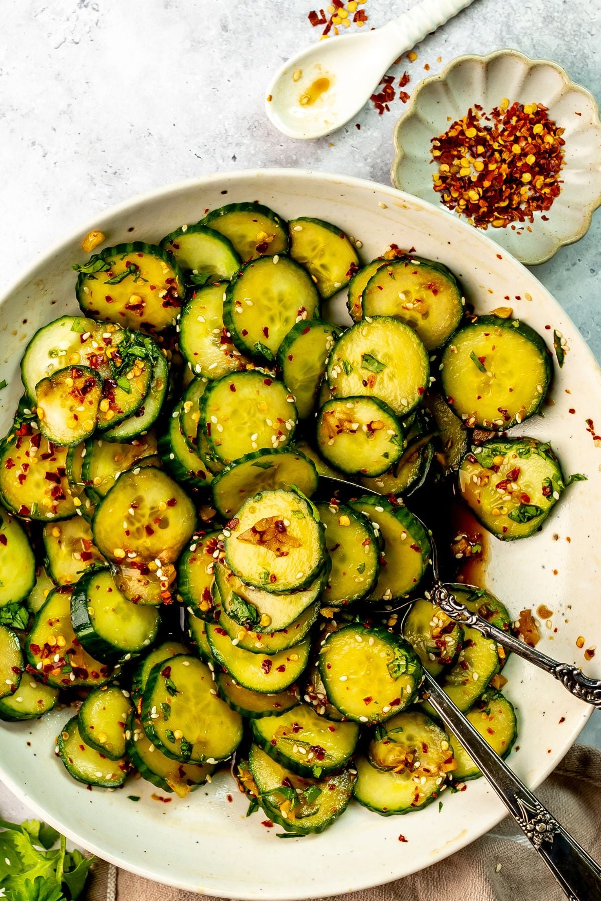 A serving spoon scoops some Asian spicy cucumber salad from a white bowl on a countertop. A small bowl of red pepper flakes sits off to the side.