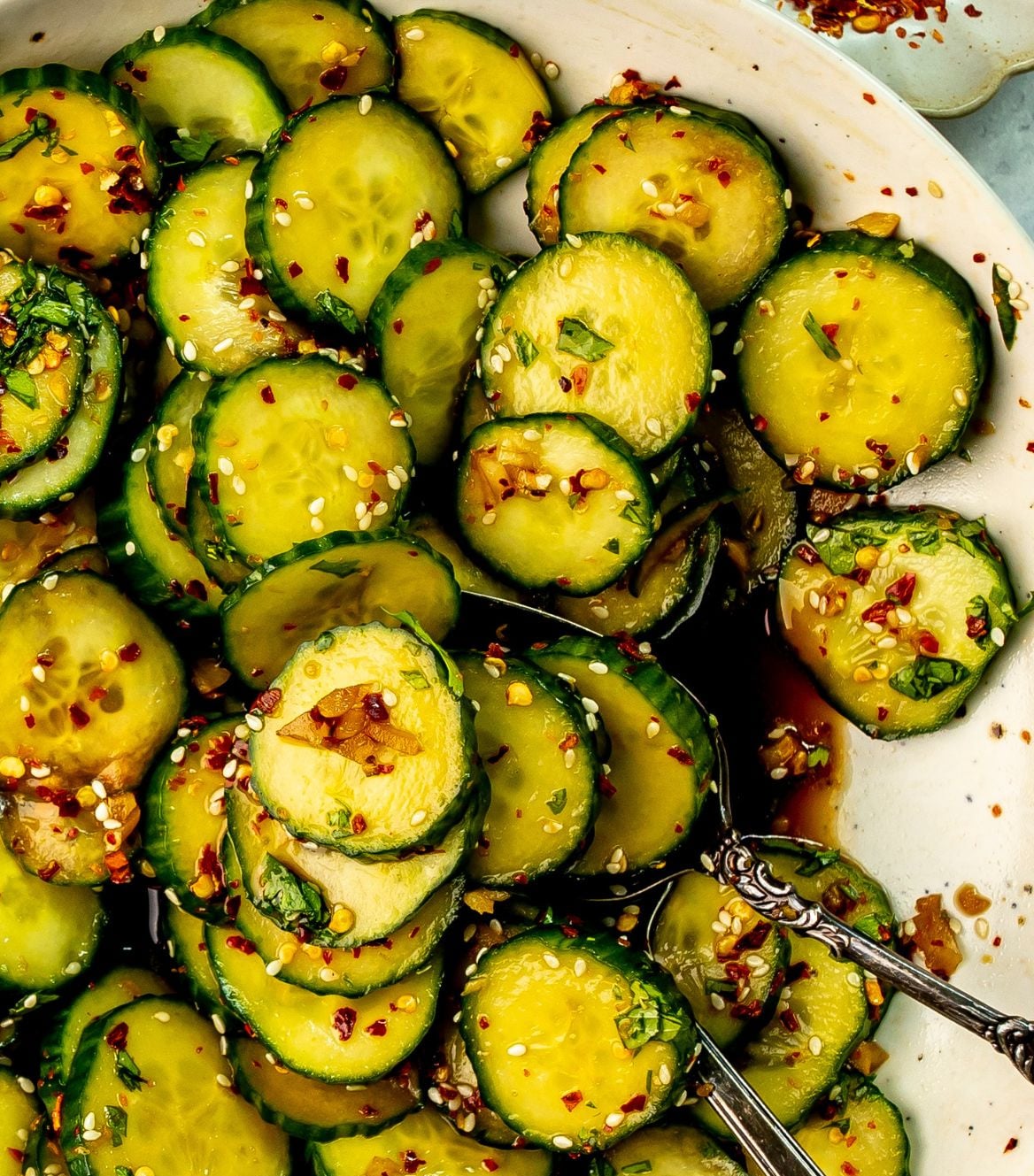 The asian spicy cucumber salad is mixed together in a white bowl with a serving spoon in it.