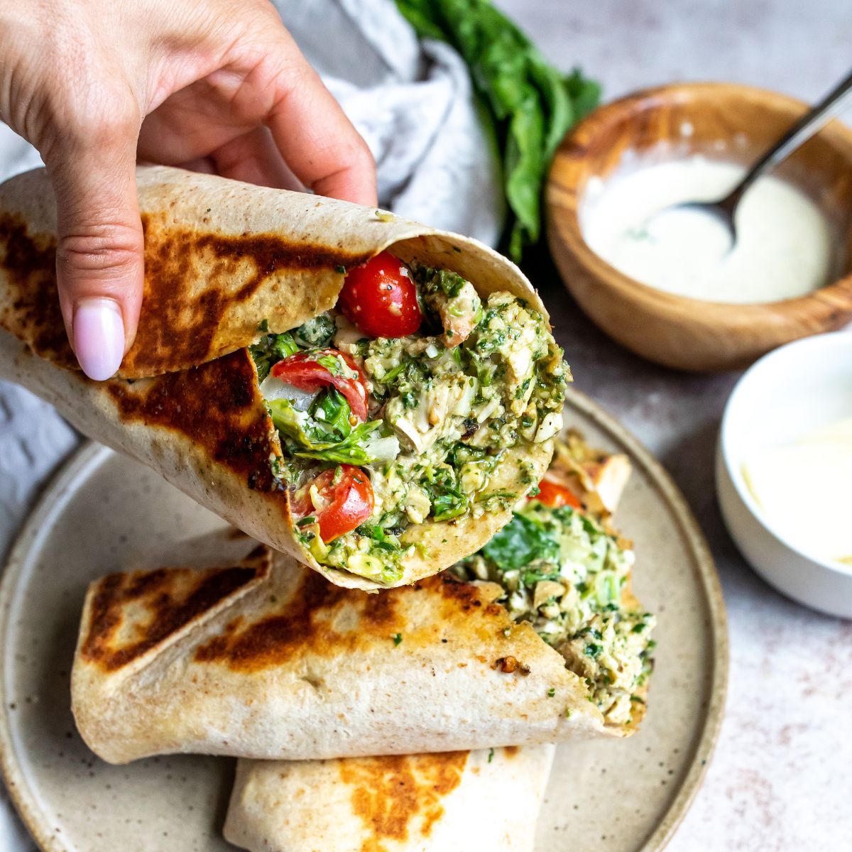 Pesto chicken wrap with pesto dressing and a hand holding the wrap, dressing on the side. 