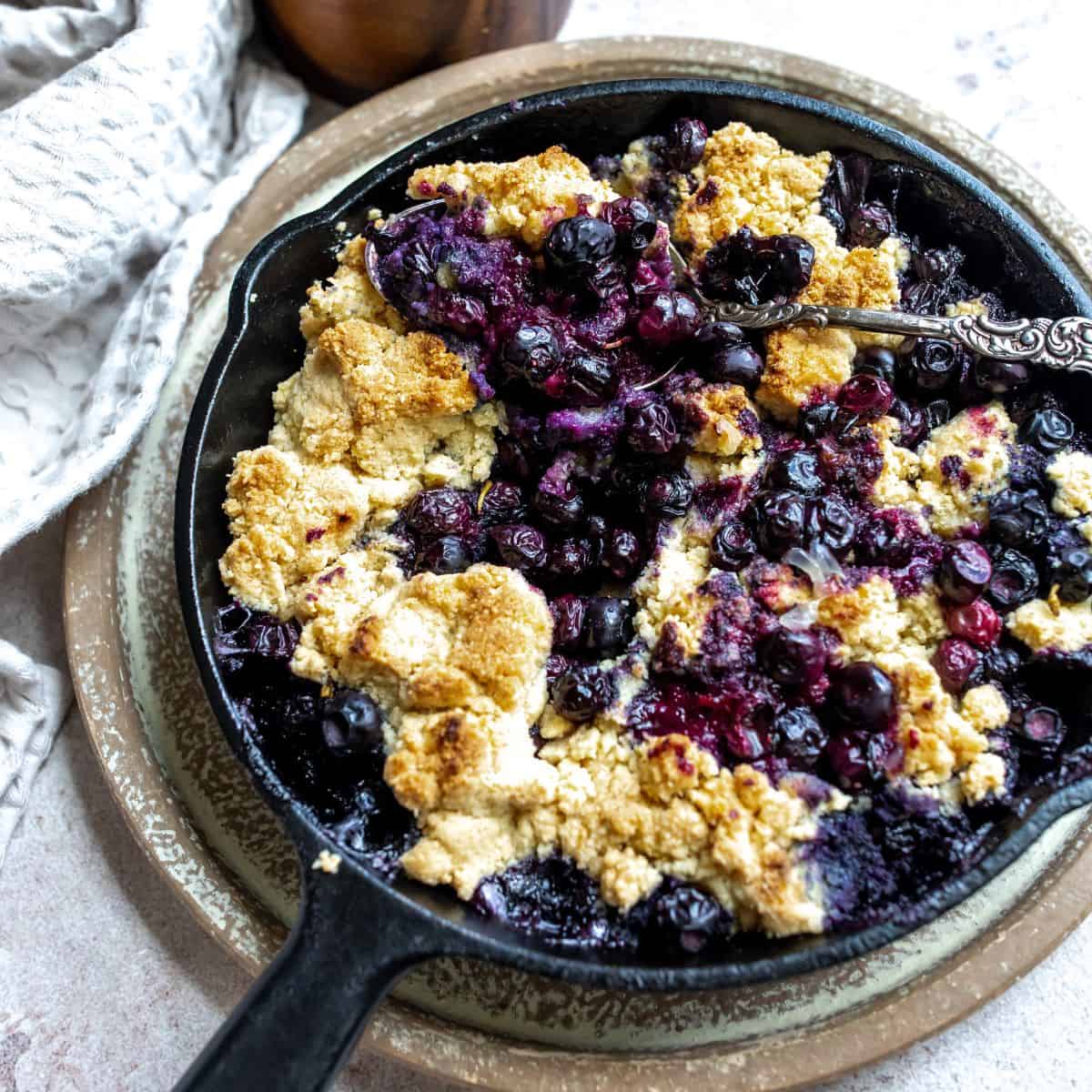 Black skillet with blueberry cobbler and a silver spoon in it. 