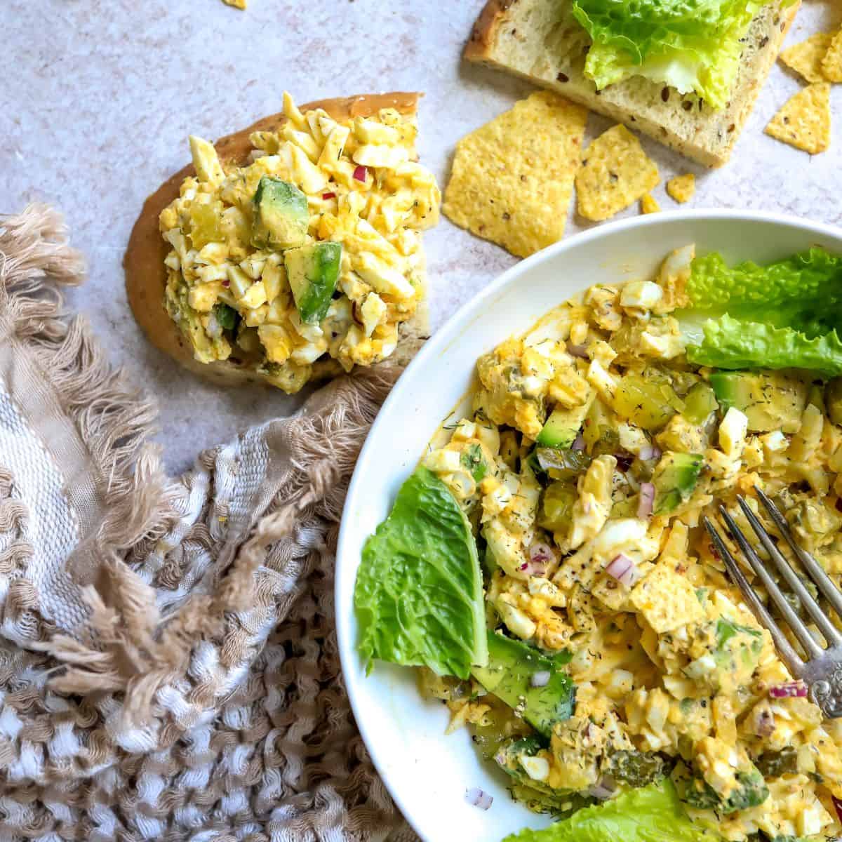 Bread with lettuce and egg mix on it. White bowl off to the side. 