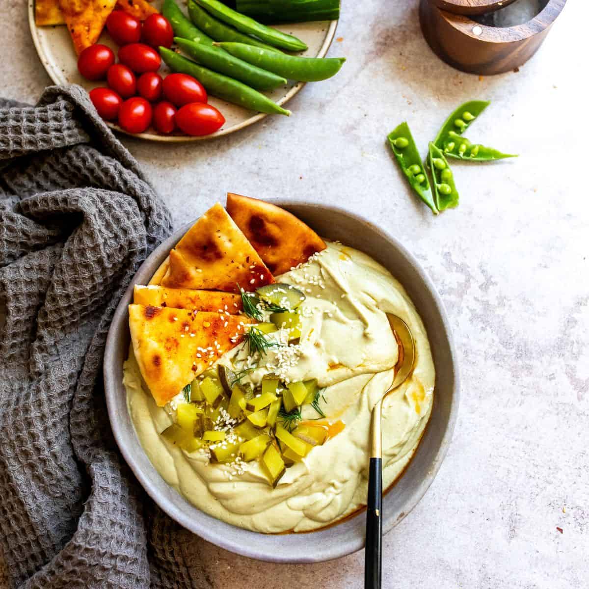 A bowl of dill pickle hummus on the table with pita chips on the side and a bowl of veggies in the background.