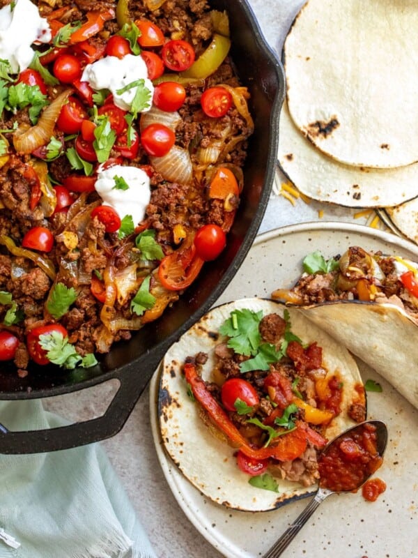 Large black skillet with fajita mix in it, topped with sour cream and tortillas on the side.