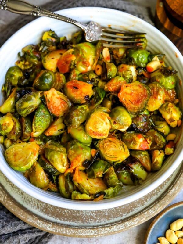 White bowl filled with brussel sprouts and sauce and a silver spoon with sauce. Peanuts on the side.