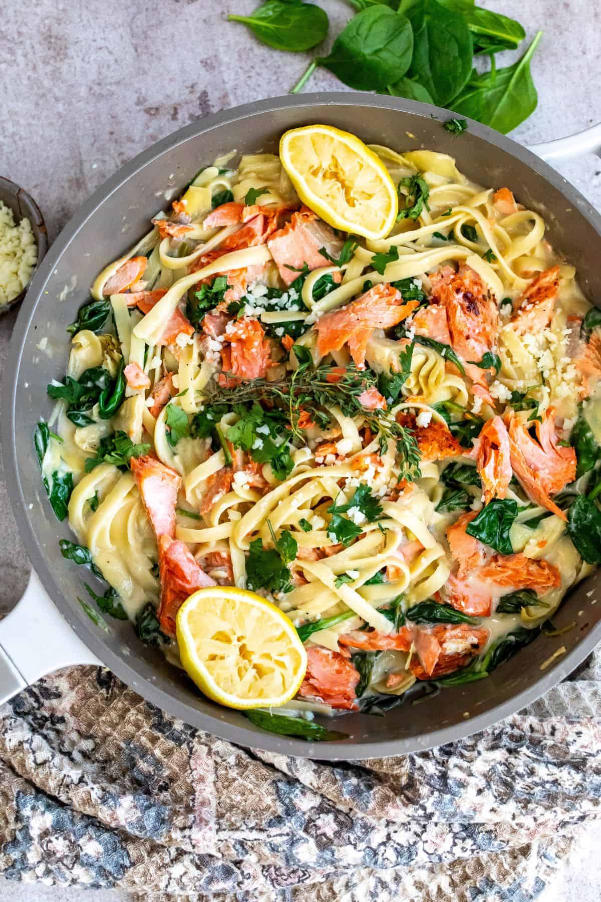 Grey skillet with pasta and cream sauce. Lemons and salmon on top. 