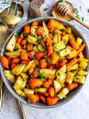 Grey pottery bowl with roasted carrots and parsnips and honey on the side.