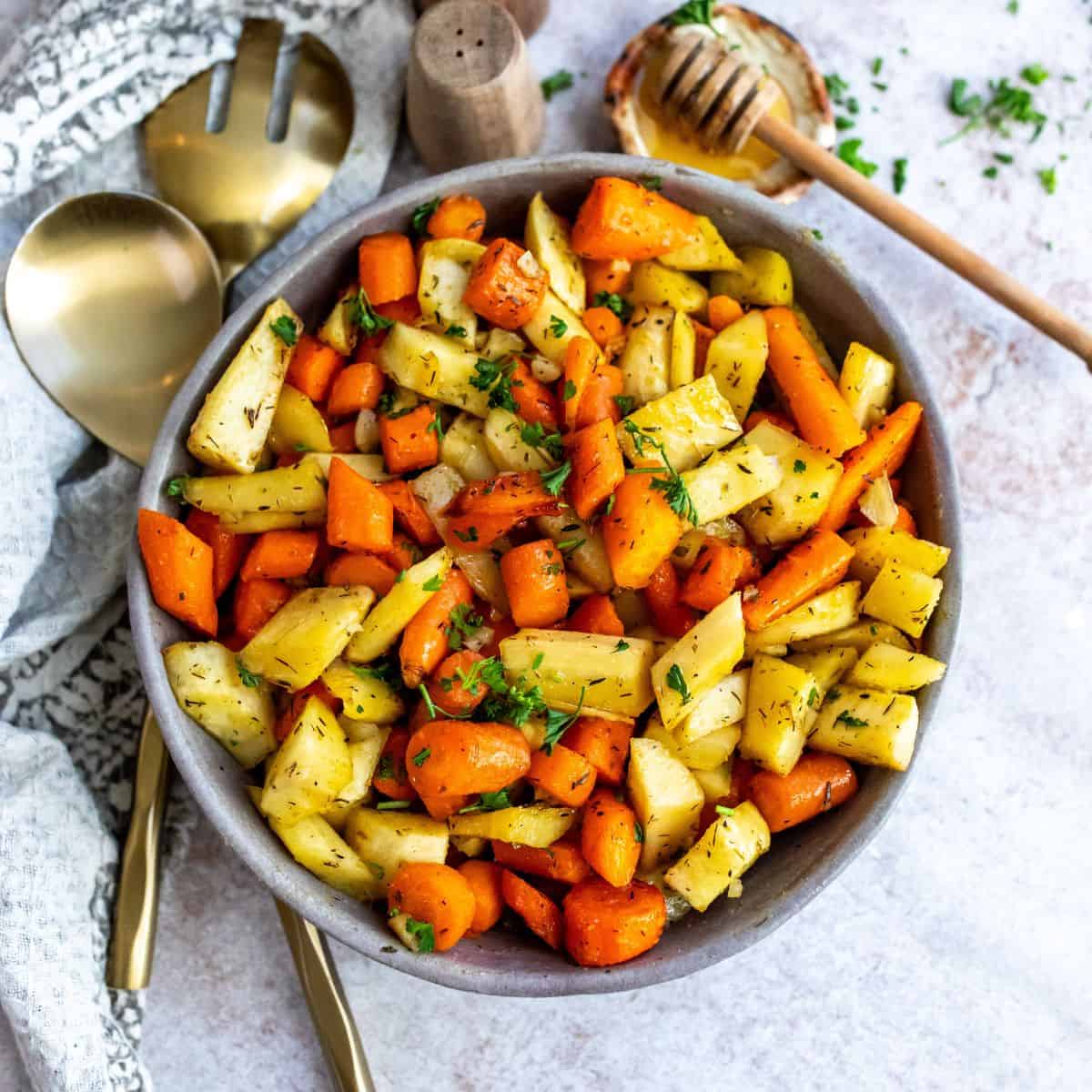 Grey pottery bowl with roasted carrots and parsnips and honey on the side.