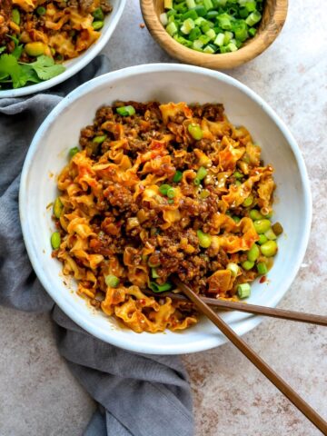 White ceramic bowl with saucy noodles and garlic. Topped with green onions and chopsticks on the side.