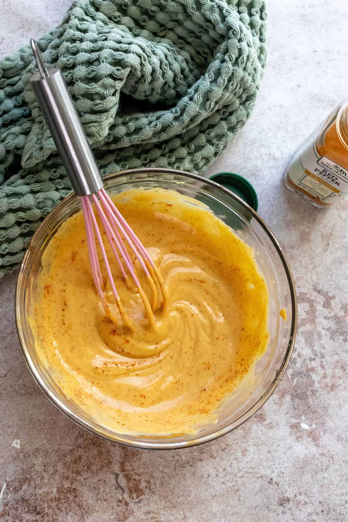 Glass bowl with honey mustard and spices mixed in and a pink whisk. 