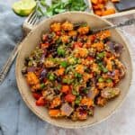 Brown ceramic bowl with sweet potatoes and quinoa, topped with cilantro and seeds. Lime and a silver fork on the side.