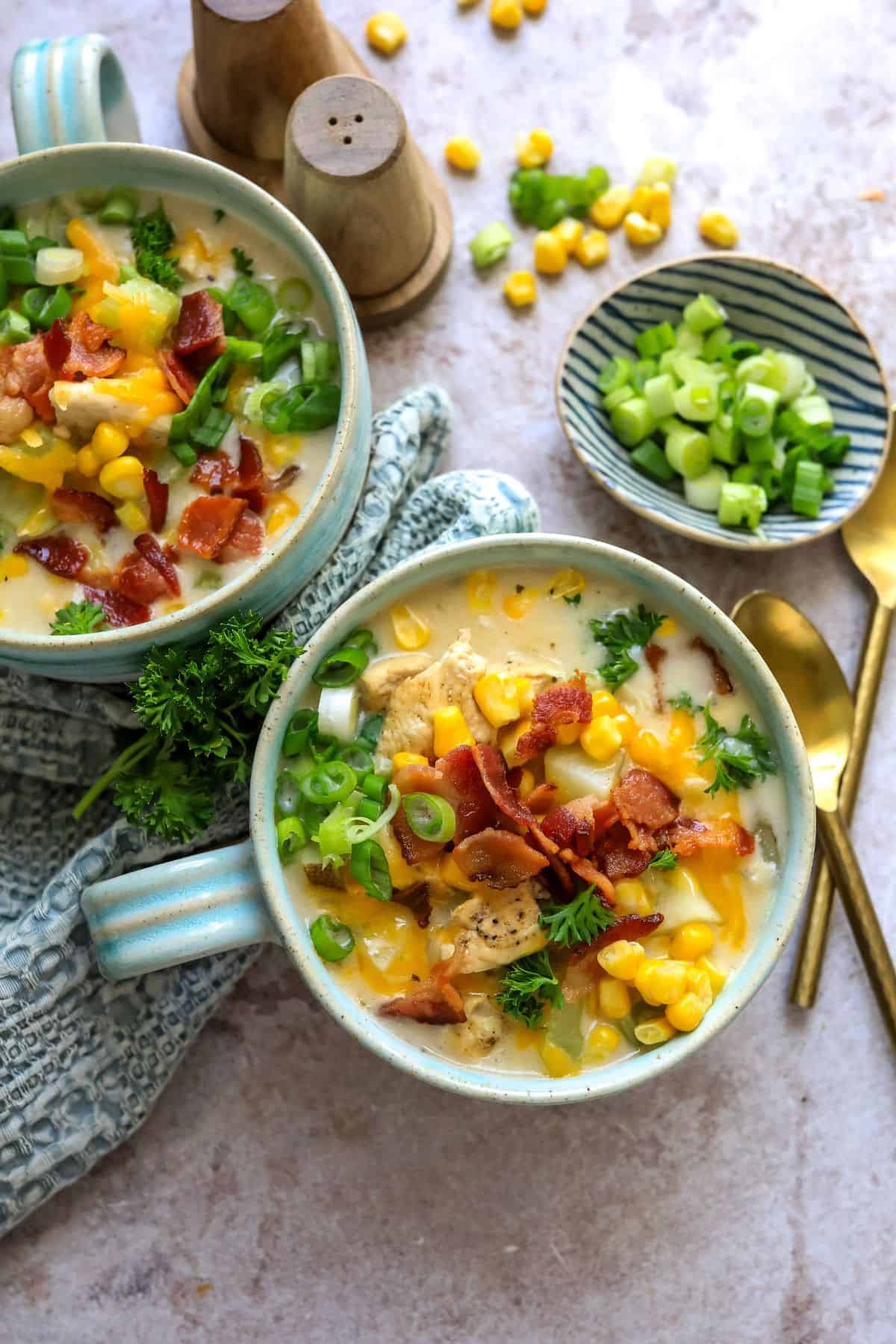 Two soup mugs with a handle on a counter with corn chowder and bacon on top. Gold spoon to the side.