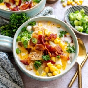 Soup mugs with a handle on a counter with corn chowder and bacon on top. Gold spoon to the side.