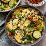 Oven safe bowls with Zucchini baked and a cheesy melt on top. A side of red pepper flakes.