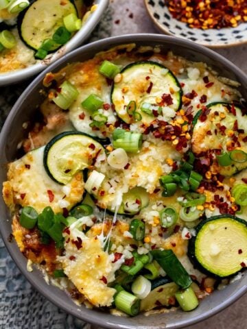 Oven safe bowls with Zucchini baked and a cheesy melt on top. A side of red pepper flakes.