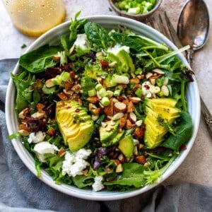White bowl with mixed greens and avocado on top. Mixing utensils to the side.