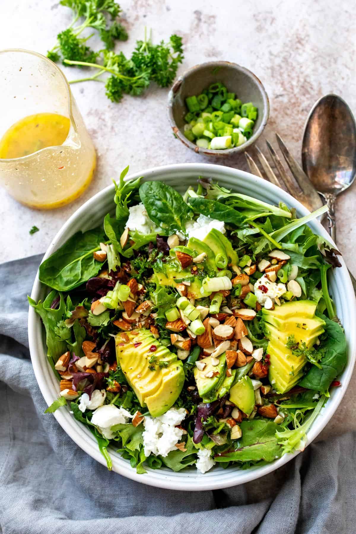 White bowl with mixed greens and avocado on top. Mixing utensils to the side. 