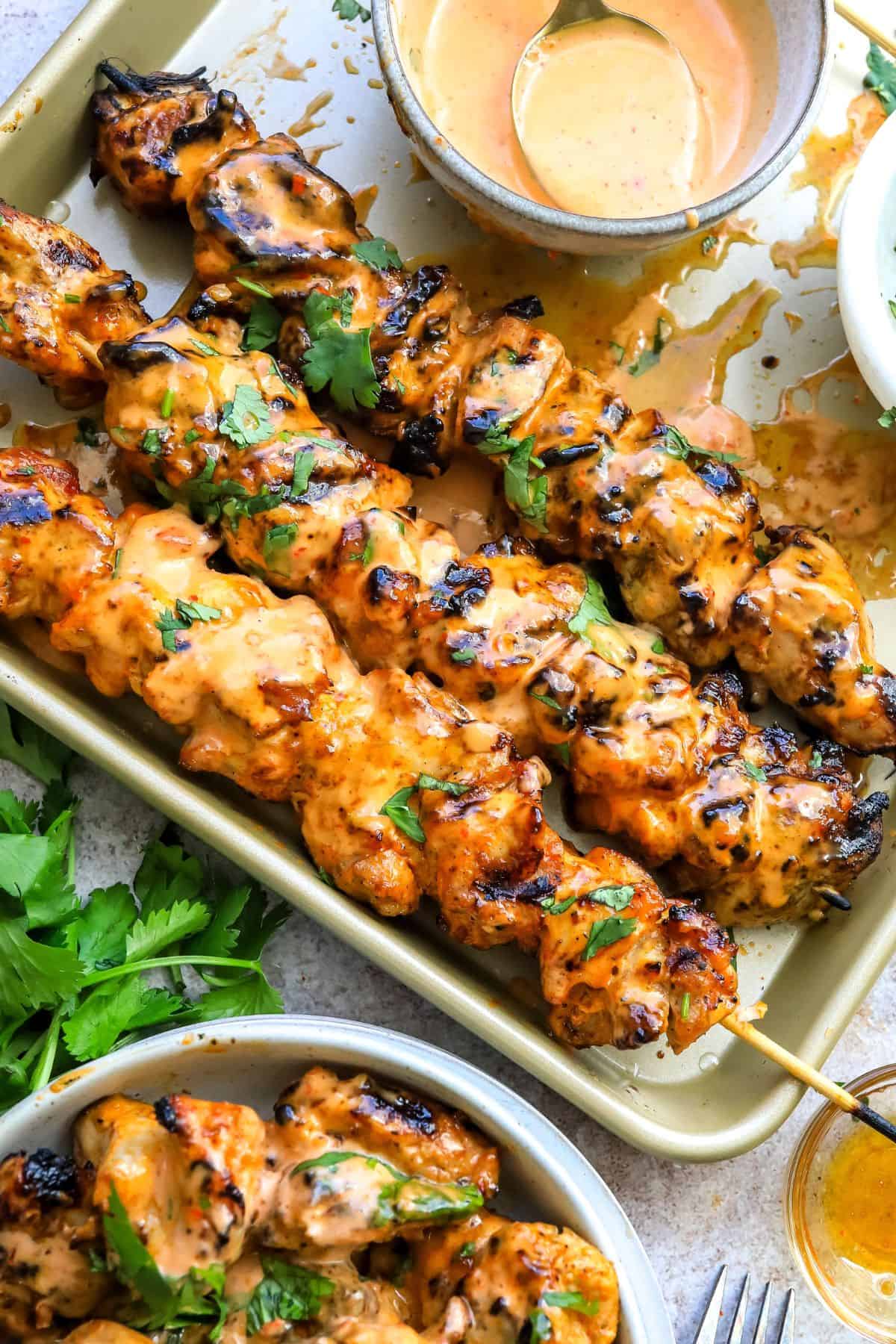 Baking sheet with spicy bang bang chicken skewers and a bowl on the side with air fried chicken skewers. 