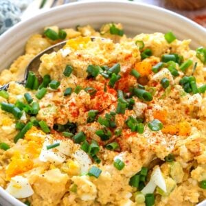 White ceramic bowl with deviled egg potato salad. Green onions on top and to the side with a spoon.