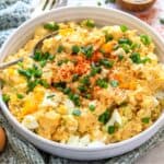 White ceramic bowl with deviled egg potato salad. Green onions on top and to the side with a spoon. Up close photo.