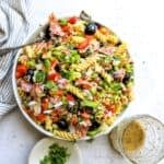 White ceramic bowl with zesty pasta salad in it with Italian dressing on the side. Bowls to the side.