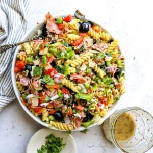 White ceramic bowl with zesty pasta salad in it with Italian dressing on the side. Bowls to the side.