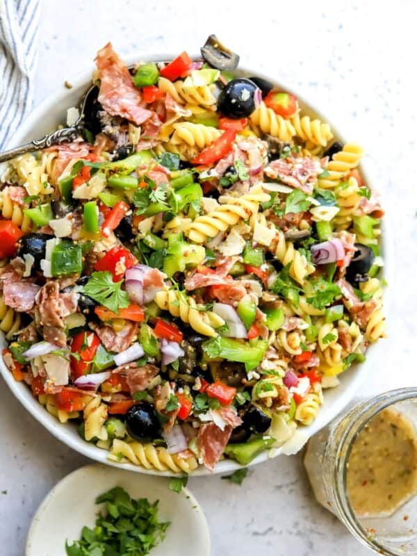 White ceramic bowl with zesty pasta salad in it with Italian dressing on the side. Bowls to the side.