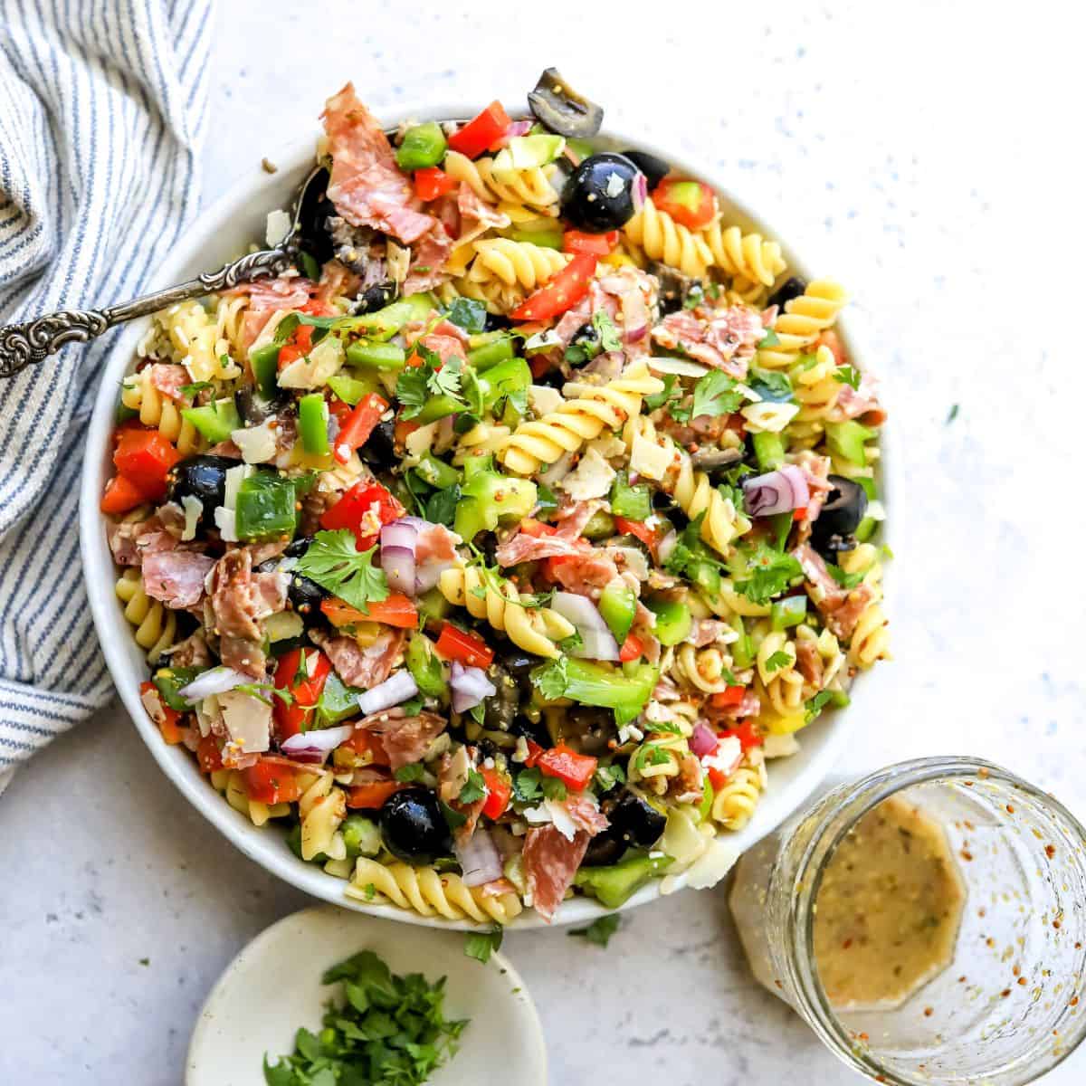 White ceramic bowl with zesty pasta salad in it with Italian dressing on the side. Bowls to the side.