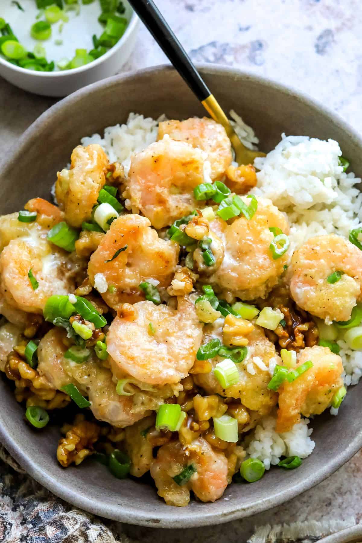 A close up image of honey walnut shrimp in a bowl over rice, garnished with green onions and candied walnuts.