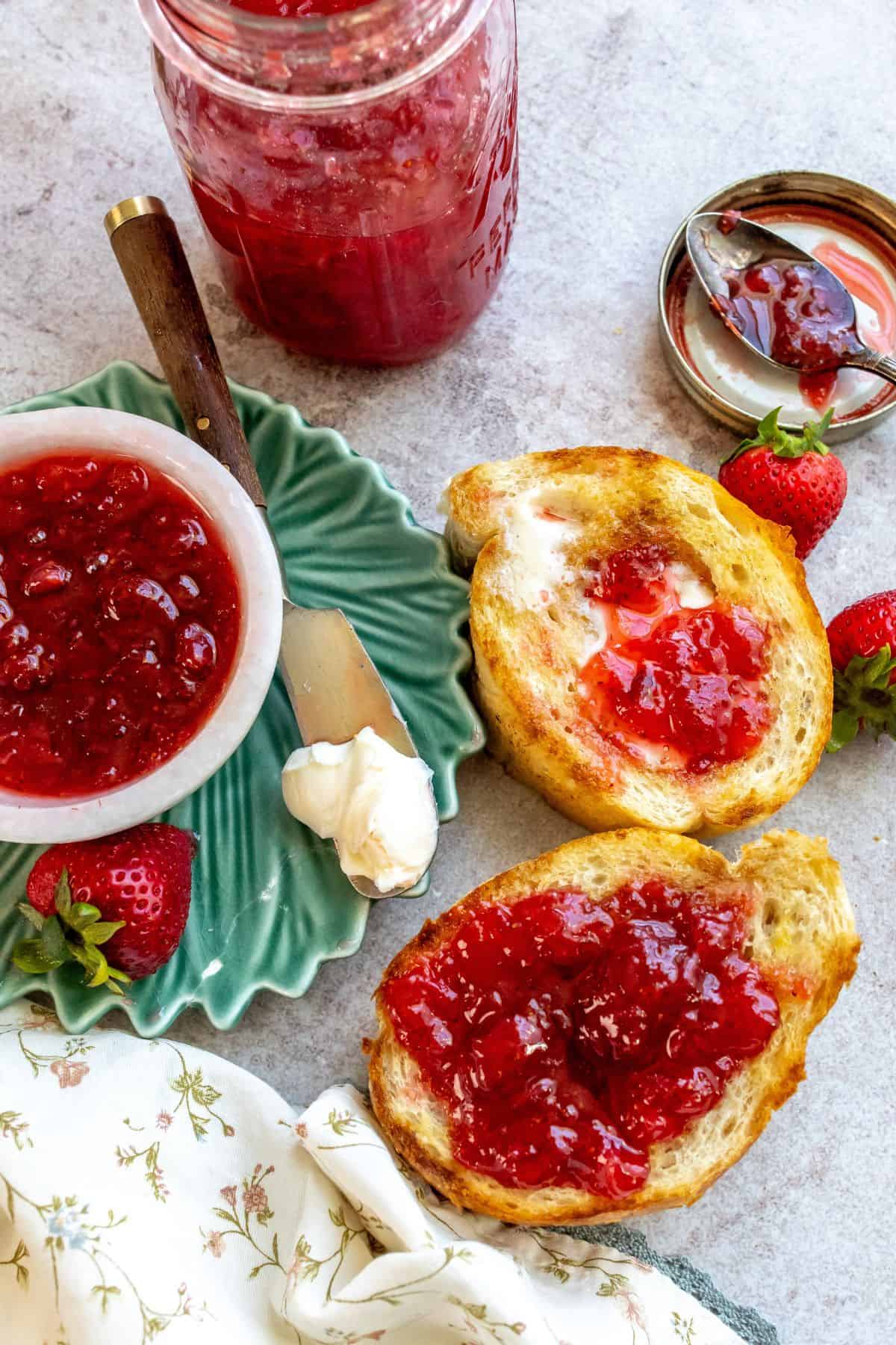 An overhead image of strawberry jam spread on two pieces of toast, with jam in a jar beside it. 