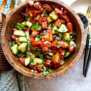 Wooden bowl with tomatoes and cucumber in it, red pepper flakes on the side.