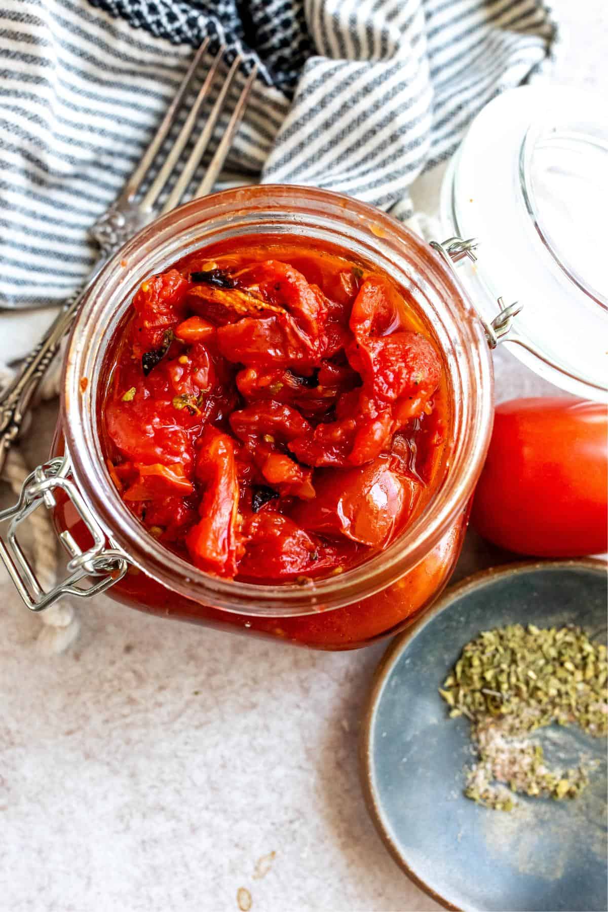 Large jar with fire roasted tomatoes in it. A fork and dried herbs to the side. 