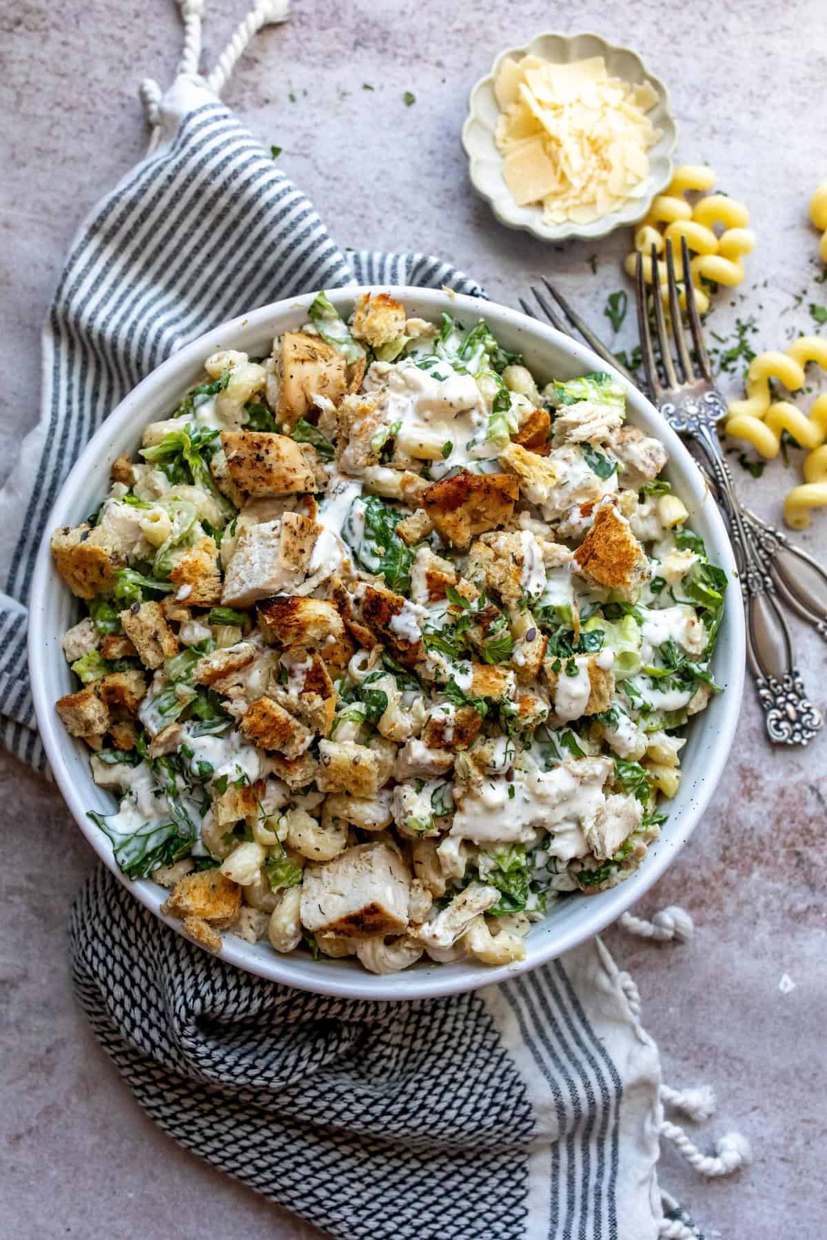 White ceramic bowl with Chicken Caesar Pasta Salad and utensils to the side. 