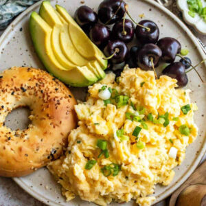 Cottage cheese scrambled eggs on a plate with a bagel, sliced avocado, and cherries.